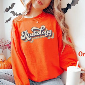 May include: Bright orange sweatshirt with the word "Radiology" in a retro font. The text is layered with blue, tan, and orange colors. The sweatshirt is long-sleeved and has a crew neck. A white mug is held in the hand.