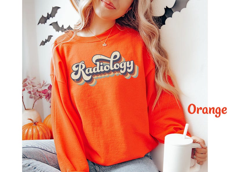 May include: Bright orange sweatshirt with the word "Radiology" in a retro font. The text is layered with blue, tan, and orange colors. The sweatshirt is long-sleeved and has a crew neck. A white mug is held in the hand.