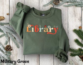 May include: Military green sweatshirt with the word "Library" in a retro font, decorated with flowers and a ladybug. The sweatshirt has long sleeves and a crew neck. The text "Military Green" is at the bottom of the image.