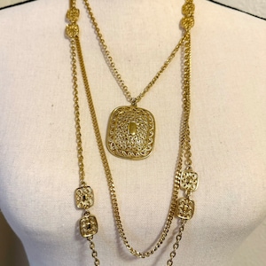 May include: A gold-toned layered necklace with a large, ornate pendant and smaller square charms. The necklace is made of multiple chains and features a delicate, intricate design.