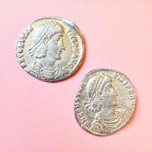May include: Two silver-colored coins with detailed profiles of a person wearing a head covering and a draped garment. The coins have inscriptions around the edges and are set against a pink background. The coins appear to be antique or vintage.