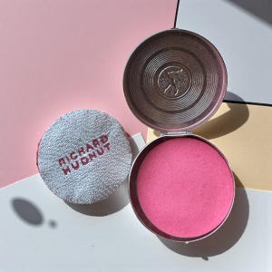 May include: Open silver compact with bright pink blush. A white powder puff with the text "RICHARD HUDNUT" is next to the compact. The compact lid has a decorative design. The background is a mix of pink, white, and yellow.