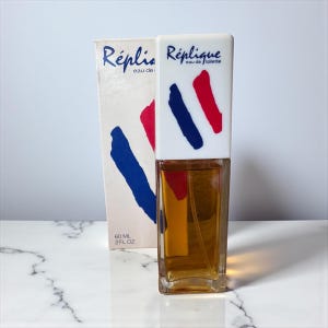 May include: A vintage perfume bottle and box with the name "Réplique". The bottle has a white cap with red and blue brushstrokes. The box has the same design and the text "eau de toilette". The bottle contains 2 fl oz of perfume.