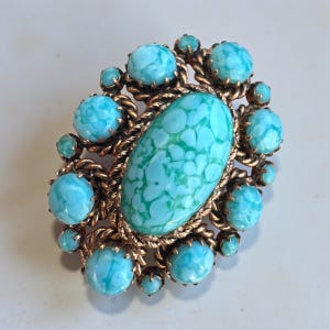 Puede incluir: Broche ovalado vintage con una piedra central turquesa y esferas turquesas más pequeñas. La broche tiene un borde dorado en forma de cuerda. La piedra central tiene un patrón moteado único. Una pieza de joyería clásica.