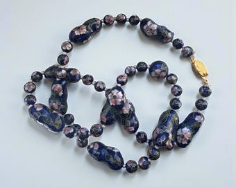 May include: A vintage necklace featuring dark blue enamel beads with a floral design in pink and white. The necklace has a gold-colored clasp and is strung on a light-colored cord. The beads are a mix of round and oblong shapes.