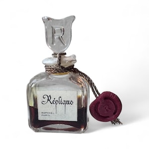 May include: A vintage perfume bottle with a clear glass body and a decorative stopper featuring the letter "R". The bottle is labeled "Réplique Raphael Paris" and has a dark liquid inside. A burgundy seal hangs from the bottle.