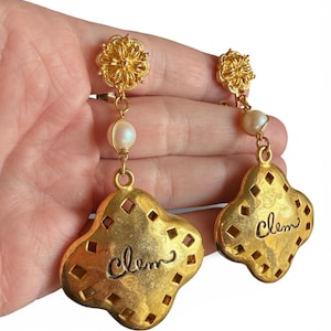 May include: A pair of gold-toned earrings. Each earring features a floral stud, a dangling pearl, and a large, quatrefoil-shaped pendant with a diamond-cut pattern and the word "Clem" in cursive. The earrings are vintage.