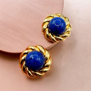 May include: A pair of gold-coloured clip-on earrings. Each earring has a deep blue, spherical stone set within a twisted gold frame. The earrings are displayed on a light pink surface.