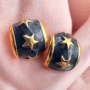 May include: A pair of vintage clip-on earrings. Each earring features a dark navy blue enamel base with gold-colored star accents and gold-colored trim. The earrings are set against a soft pink background.