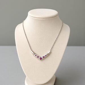 Puede incluir: Un collar en forma de V de tono plateado con un colgante central en chevron engastado con piedras redondas rojas y blancas transparentes alternadas. El colgante está unido a una delicada cadena plateada, creando un diseño estructurado.