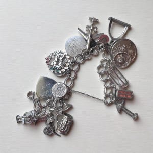 May include: A vintage silver charm bracelet featuring a variety of charms, such as a plane, a medal, a beer stein, and a shield. The charms are linked to a chain bracelet. The bracelet is on a white surface.