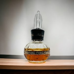 May include: A vintage glass perfume bottle with a clear glass stopper and a black and gold decorative band around the middle. The bottle is filled with a light brown liquid.