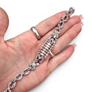 May include: A silver-toned bracelet with clear, sparkling rhinestones. The bracelet features a series of oval links and a central section with rectangular and round stones. The clasp is visible at the end of the bracelet.