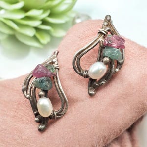 May include: A pair of silver-toned earrings with a leaf-like design. Each earring features a cluster of gemstones in pink and green hues, along with a white pearl. The earrings are displayed on a pink fabric.