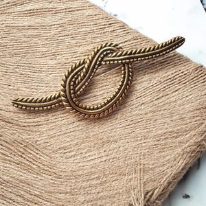 May include: A gold-toned metal brooch in the shape of a knot. The knot is detailed with a braided rope design.