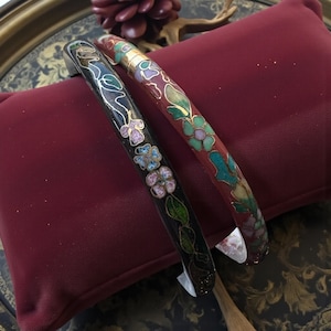 May include: Two decorative enamel bangles. One is black with floral designs in pink, blue, and green. The other is red with gold accents and floral designs in green, purple, and teal. Both are resting on a red pillow.