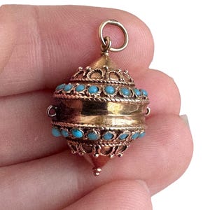 May include: A gold-colored pendant with a rounded shape, adorned with small turquoise-colored stones. The pendant has intricate filigree detailing and a loop at the top for hanging. The design is reminiscent of vintage or antique jewelry.