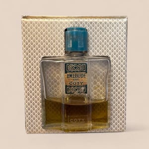 May include: A vintage perfume bottle of Emeraude de Coty, with a teal cap and a rectangular glass bottle. The bottle is set inside a decorative box with a repeating leaf pattern. The perfume liquid is amber-colored.