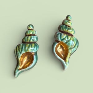 May include: Two ceramic seashell sculptures. The shells are a mix of green and yellow with a gold interior. The spiral design is detailed with brown accents. The background is a solid light green.
