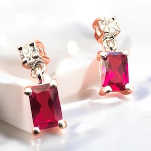 May include: A pair of rose gold earrings with a rectangular, deep red gemstone and a small, clear, square-cut stone at the top. The earrings are set against a white background, highlighting the jewelry's sparkle and design.
