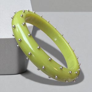 May include: A light green bangle bracelet with small, silver-colored studs evenly spaced around the exterior. The bracelet has a smooth, glossy finish and is displayed on a neutral background. This piece of jewelry is a stylish accessory.