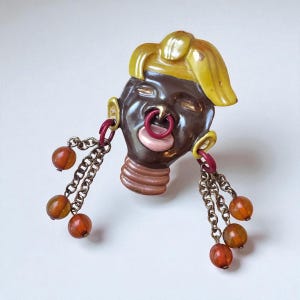 May include: A vintage brooch in the shape of a stylized face with dark brown skin, a yellow hairstyle, and a pink tongue. The brooch has dangling chains with orange beads and red accents.