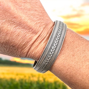 May include: A silver bracelet with a braided design. The bracelet is made of multiple strands of silver wire that are woven together to create a complex pattern.