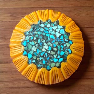 May include: A round, decorative object with a vibrant design. The outer edge is a warm yellow with carved vertical lines. The center is filled with turquoise and teal-colored stones, creating a mosaic effect. The object is set against a wooden background.