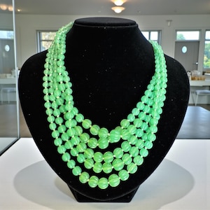 Puede incluir: Collar de varias hebras con cuentas verdes claras y acanaladas. El collar se exhibe en un busto de maniquí negro. Las cuentas están ensartadas con hilo blanco, creando un efecto en capas. El collar es una pieza llamativa.