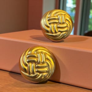 May include: Two gold-colored, round cabinet knobs with a woven design. The knobs are made of metal and have a shiny finish. They are sitting on a peach-colored surface.
