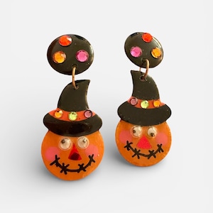 Peut inclure: Une paire de boucles d'oreilles sur le thème d'Halloween. Chaque boucle d'oreille représente un visage de citrouille orange avec un sourire cousu et un chapeau de sorcière noir. Les boucles d'oreilles sont ornées de strass colorés.