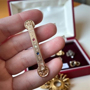 May include: A gold-colored bar pin with a series of colorful gemstones. The pin features a rectangular design with decorative engravings and circular accents at each end. The gemstones are red, white, and blue.