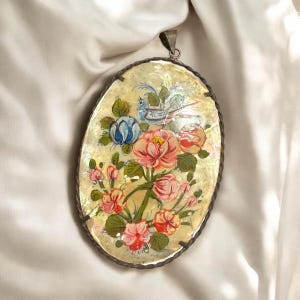 Pode incluir: Pingente oval com um desenho floral pintado à mão. O pingente apresenta flores rosa, vermelhas e azuis com folhas verdes e um pequeno pássaro azul. O pingente tem uma moldura de metal prateado e uma argola para pendurar.