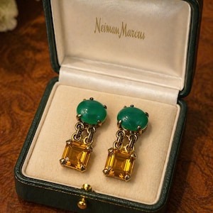 May include: A pair of vintage earrings in a green leather box. Each earring features a green oval stone at the top, connected by a gold chain to a square, amber-colored stone. The box is lined with cream-colored fabric and has the words "Neiman Marcus" printed on the inside.
