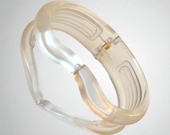 Könnte beinhalten: Ein klarer, durchscheinender Armreif mit einem einzigartigen, ineinandergreifenden Design. Der Armreif hat ein Scharnier mit einer kleinen goldfarbenen Metallkomponente. Die Gesamtästhetik ist modern und minimalistisch, mit Fokus auf dem Zusammenspiel von Licht und Form.