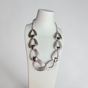 Può includere: Una collana statement color argento caratterizzata da una serie di maglie testurizzate a forma di goccia interconnesse che formano un anello continuo sul fondo. Il metallo ha una finitura spazzolata con accenti lucidi sulle giunture.