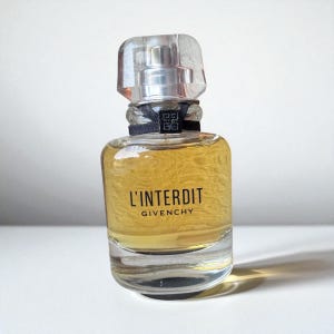 Op de afbeelding: Een heldere glazen parfumfles met een zilveren verstuiver en een transparante, rechthoekige dop. De fles bevat een goudkleurige vloeistof en is voorzien van het opschrift "L'INTERDIT GIVENCHY" in zwarte tekst. Een zwart lint is om de hals van de fles gewikkeld.