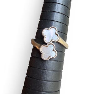 May include: A silver-toned ring with two clover-shaped mother-of-pearl inlays. The ring is displayed on a black ring sizer. The ring's design features a delicate, openwork style, with the clover shapes set against a silver band.