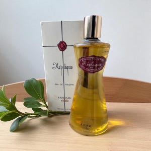 May include: A vintage bottle of Réplique Eau de Toilette perfume by Raphael Paris. The clear glass bottle has a unique hourglass shape with a gold cap. The packaging includes a white box with the perfume's name and logo.