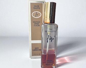 May include: A clear glass bottle of "Fidji" Eau de Toilette by Guy Laroche, with a gold cap and a light brown liquid. The bottle is rectangular with the "Fidji" logo and "Eau de Toilette" printed on it. The box is beige with the same logo.