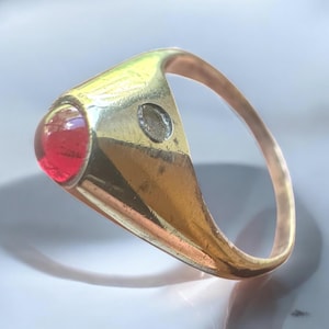 May include: A gold-toned ring with a large, round, red gemstone. The ring has a classic design with a smooth band and a setting that holds the gemstone securely. The gemstone is a vibrant red color, and the ring appears to be well-crafted.