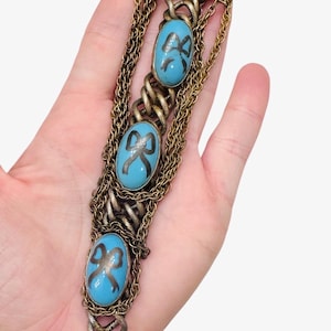 May include: Vintage bracelet featuring oval turquoise-colored stones with black bow designs, set in ornate gold-toned metal. The bracelet has multiple chains and a decorative gold-colored teardrop-shaped charm at the end. A unique piece of jewelry.