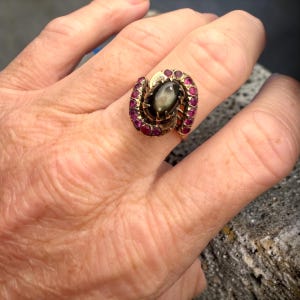 May include: A gold ring featuring a central oval black star sapphire cabochon surrounded by a curved, bypass-style setting adorned with a row of small, round, vibrant pink rubies set in a polished yellow gold band with a textured finish.