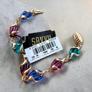 May include: A gold-tone link bracelet featuring alternating translucent blue, purple, and teal oval beads encased in gold metal swirls. A black retail tag with the text SAVVY and a barcode is attached to the bracelet, which ends in a ribbed gold clasp.