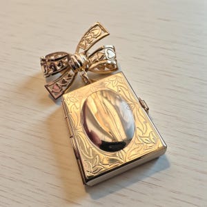 May include: A gold-colored locket with a bow-shaped clasp. The rectangular locket has an oval center and is decorated with floral patterns. The bow clasp features heart-shaped cutouts.