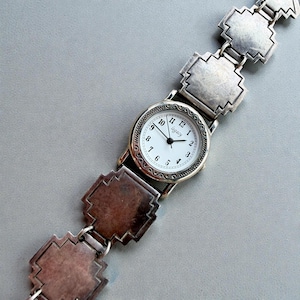 Könnte beinhalten: Eine silberfarbene Legacy-Uhr mit weißem Zifferblatt und schwarzen Ziffern. Die Uhr hat eine dekorative Lünette und ist an einem silbernen Armband mit geometrischen Gliedern befestigt. Die Uhr liegt auf einer grauen Oberfläche.