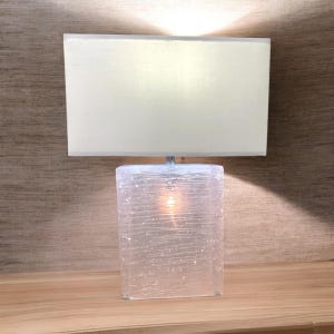 May include: A white lampshade with a clear glass base with a textured surface. The lamp is turned on and the light is shining through the glass.