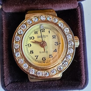 May include: A gold-toned ring watch with a round face and a bezel encrusted with clear crystals. The watch face is cream-colored with black numerals and hands. The word "DIGITS" is printed above the center.