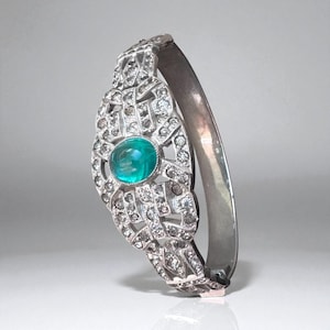 May include: A silver-toned hinged bracelet with an ornate, openwork design. The focal point is a large, round, teal-colored gemstone, surrounded by smaller, sparkling stones. The bracelet is a classic piece of jewelry.