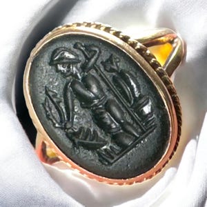 May include: A gold ring with an oval, dark green stone. The stone is carved with a detailed relief of two figures, one holding a trident. The ring has a twisted rope design around the stone and a gold band.